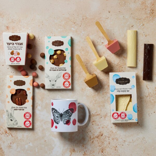 מערך מוצרי השוקולד משתה המלך כולל מגוון פריטים המסודרים על משטח בעל מרקם, כולל חטיפי שוקולד ושוקולדים בקופסאות. ספל בעיצוב פרפר משלים את הקולקציה, עם אריזות צבעוניות המציגות נושאים של בעלי חיים.