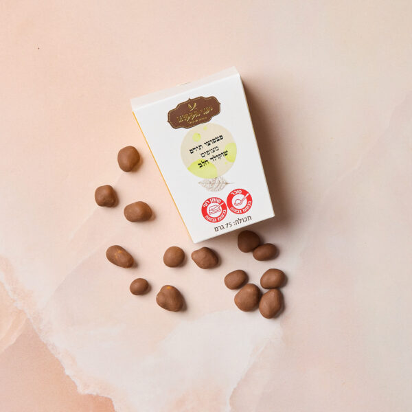 קופסה לבנה עם הכיתוב "IMG_2095" וטקסט בעברית ("תירס קלוי מצופה שוקולד חלב") וסמלי אישור מונחת על משטח שיש ורוד, מוקפת באגוזים או סוכריות מצופים שוקולד.