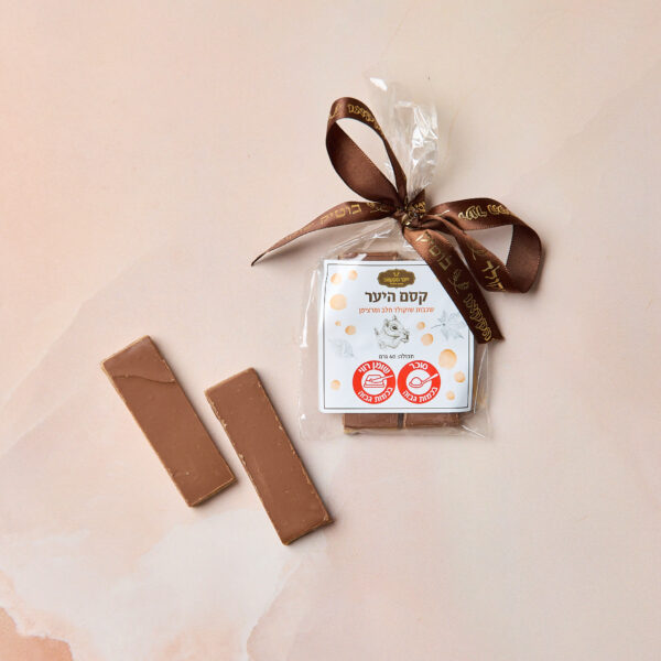 שקית מתנה שקופה עם סרט חום מכילה את IMG_2103 — חבילת שוקולד מריר עם תווית בעברית — לצד שתי חתיכות שוקולד לא עטופות על משטח שיש ורוד בהיר.