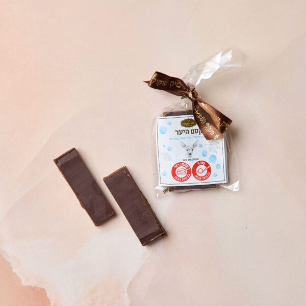 שני מלבני שוקולד מריר מונחים לצד ממתק מרציפן עטוף בסרט חום ותווית כחולה-לבנה בעברית עם שני לוגואים אדומים, והכל מונח על משטח דמוי שיש בהיר. שם המוצר: IMG_2104.
