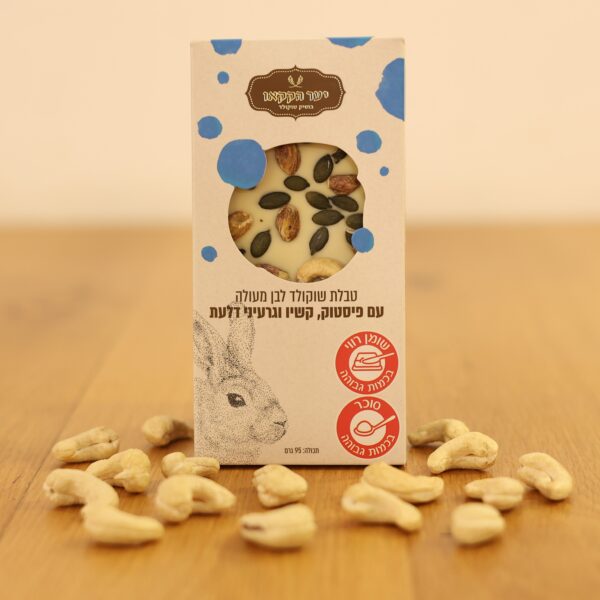 קופסה של 3I0A3102, שוקולד לבן עם פיסטוק, קשיו וגרעיני דלעת, מונחת על משטח עץ מפוזר בקשיו. על האריזה מופיע איור של ארנב וטקסט בעברית.