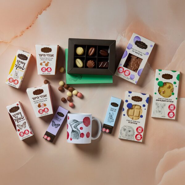 חבילת בשורות טובות כוללת מגוון פריטי שוקולד על שולחן, הכוללים שוקולדים בקופסאות וספל בעלי חיים. התצוגה מועשרת באגוזים מפוזרים בשוקולד, והאריזה מדגישה צבעים ועיצובים מגוונים.