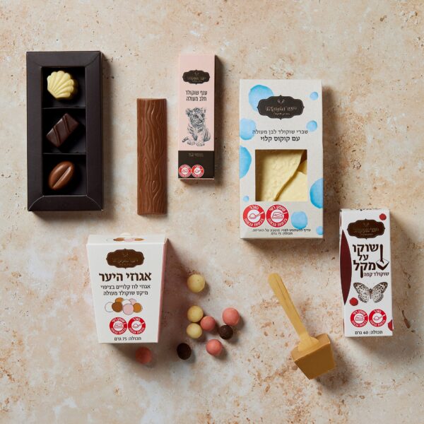 מבחר פינוקים מתוקים מוצגים על משטח שיש, הכוללים חשב עליך עם שוקולדים בקופסאות, חפיסת שוקולד, סוכריות צבעוניות וקרטון משקאות עם מטבל דבש מעץ בקרבת מקום, יחד עם פריטים באריזה המסומנת בשפה זרה.