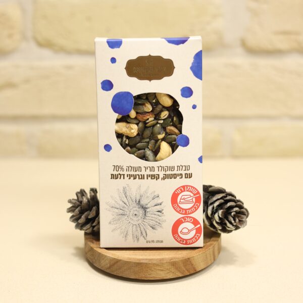 מוצר 3I0A2953, קופסה של קליפת שוקולד מריר 70% עם פיסטוקים, גרעיני דלעת וחמניות, ניצבת על מגש עץ בין שני אצטרובלים. הקופסה מציגה טקסט עברי ואיורים של גרעינים על רקע לבנים בהירות.