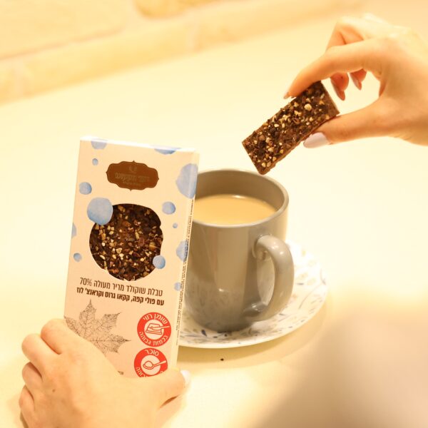 אדם טובל חטיף שוקולד מלבני 3I0A3017 עם אגוזים בקפה, כשהוא אוחז בקופסה המסומנת בעיגולים כחולים ובכיתוב בעברית. הסצנה מוצגת על משטח עבודה בהיר.