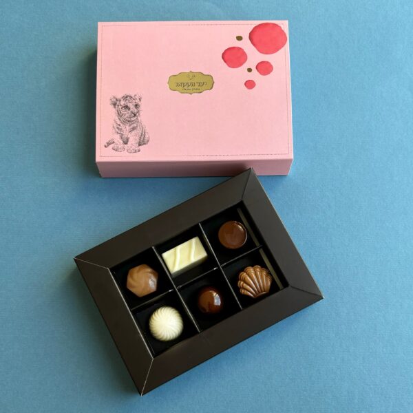 תמונה של מארז בגודל מלא עם מכסה ורוד ובו רישום של כלב ונקודות אדומות מונחת לצד מגש שחור פתוח של שישה פרלינים שוקולד שונים, כולם מוצגים על רקע כחול.