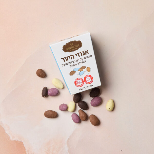 קופסה קטנה ולבנה עם הכיתוב IMG_2092 ועליה טקסט בעברית מונחת על משטח ורוד, מוקפת במיקס שקדים — מערך של שוקולד בטעמים צבעוניים ושקדים מצופים טעימים.