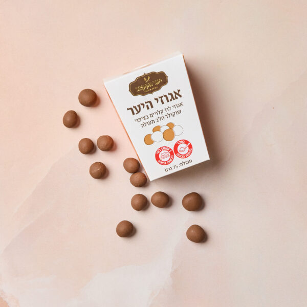 קופסה עם הכיתוב IMG_2097 בעברית ובצבעי זהב ואדום מונחת על משטח ורוד בהיר, מפוזרת אגוזי לוז מצופים שוקולד חלב. על הקופסה מופיעה איור קטן של הממתקים.