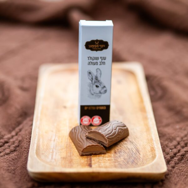 קופסה עם איור של ארנבת וטקסט בעברית ניצבת על מגש עץ, ובחזיתה שתי חתיכות של "ענף חלב חמאת בוטנים" שלא עטופות חלקית, משובצות על רקע בד חום עם טקסטורה.