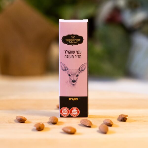 קופסה קטנה שכותרתה "ענף מריר שקדים1" עם כיתוב עברי ופרצוף של צבי ניצבת זקופה על משטח מטושטש, מוקפת בזרעים חומים או אגוזים מפוזרים, מה שמרמז על מרכיבים מבוססי שקדים או טבעוניים. הרקע מעט ערום.