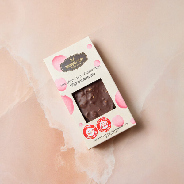 חפיסת שברים מריר עם פיסטוק שוקולד מלבנית בקופסה בצבע בז' עם עיגולים ורודים וטקסט בעברית, מוצגת על משטח שיש ורוד. חתיכות השוקולד המריר והפיסטוק נראות חלקית מבעד לחלון האריזה השקוף.