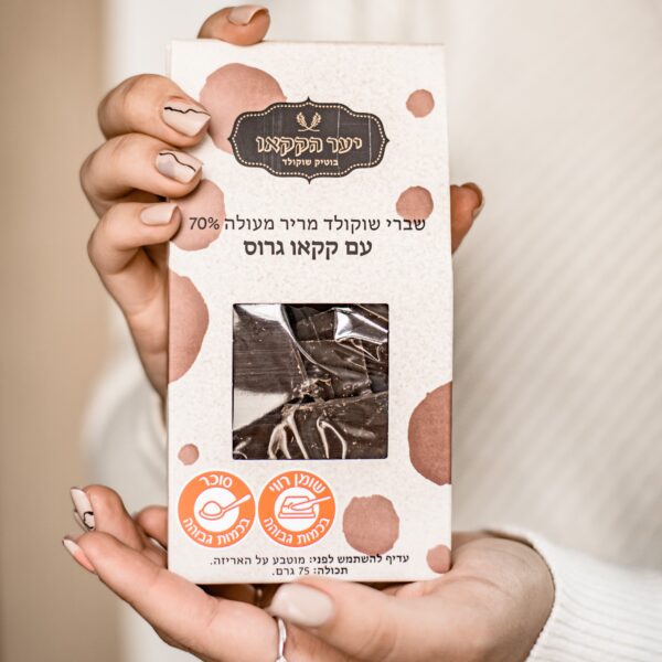 יד אוחזת בקופסה של קליפת שוקולד שברים מריר קקאו עם אגוזי לוז וניבס של קקאו. חבילת הבז' כוללת כתמים חומים, טקסט בעברית, חלון לצפייה בשוקולד, בתוספת אייקונים של "טבעוני" ו"ללא סוכר".