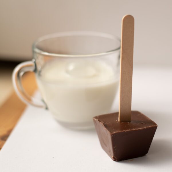 כוס חלב זכוכית שקופה מונחת ליד גוש שוקו מריר על משטח לבן.