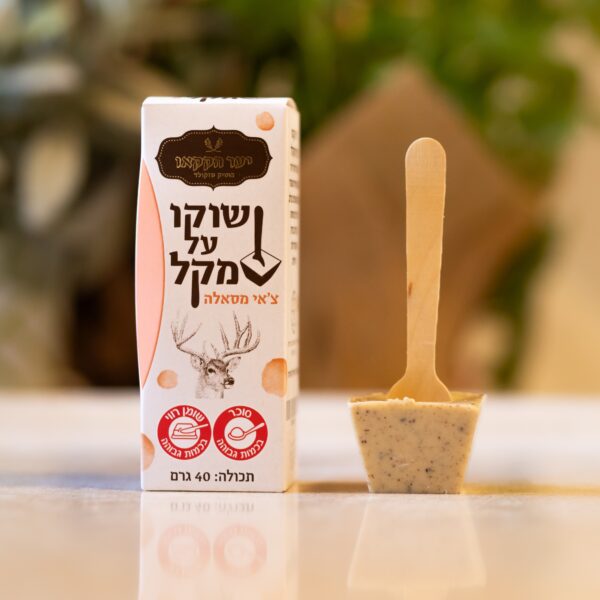 קופסה עם טקסט עברי ואיור של צבי ניצבת לצד מקל עץ ובו גוש שוקולד לבן מנומר של שוקו צאי. הרקע המטושטש עדין מתאפיין בגוונים ירוקים וחומים.