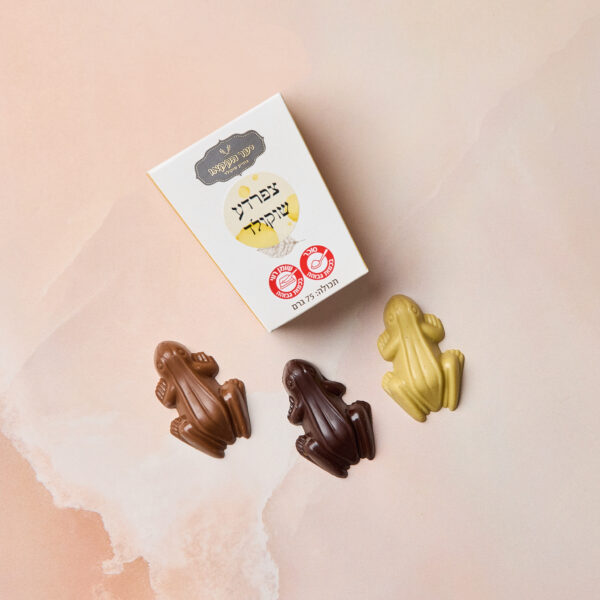 קופסה עם הכיתוב IMG_2113 וטקסט בעברית לצד שלוש צפרדעי שוקולד — חלב, מריר ולבן — על משטח שיש ורוד בהיר. כל צפרדע שוקולד מגיעה עם כרטיס אספנים קלף.