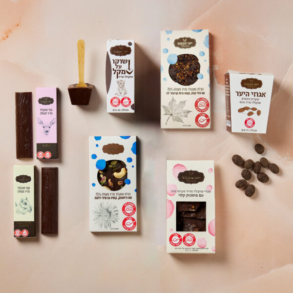 מגוון מוצרי שוקולד, ביניהם חטיפי מריר טבעוני, שוקולדים בקופסאות ואגוזים מצופים בשוקולד, מסודרים על משטח בהיר. האריזה מציגה עיצובים מגוונים וטקסט שאינו אנגלי. כף עץ נראית גם בקרבת מקום.