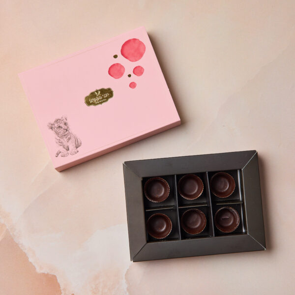 IMG_2064 מציג קופסה ורודה עם איור של נמר ומעגלים אדומים לצד מגש פתוח עם שש כוסיות שוקולד, כל אחת בתא נפרד, המוצגות על רקע רך ובהיר.