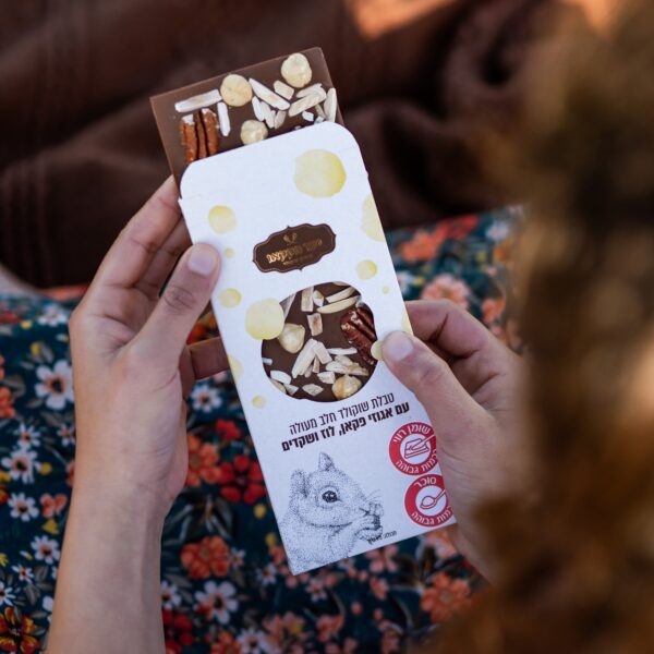 אדם מחזיק חטיף שוקולד פתוח "טבלה חלב ואגוזים", שהוצא חלקית מאריזה לבנה וצהובה עם איור של עכבר וטקסט בעברית, על רקע שמלה עם דוגמת פרחים.