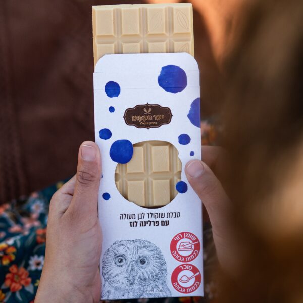 אדם מחזיק חטיף טבלה לבן לוז, עטוף חלקית באריזה לבנה עם כתמים כחולים, טקסט בעברית ואיור ינשוף. חטיף השוקולד מציג דוגמת רשת.