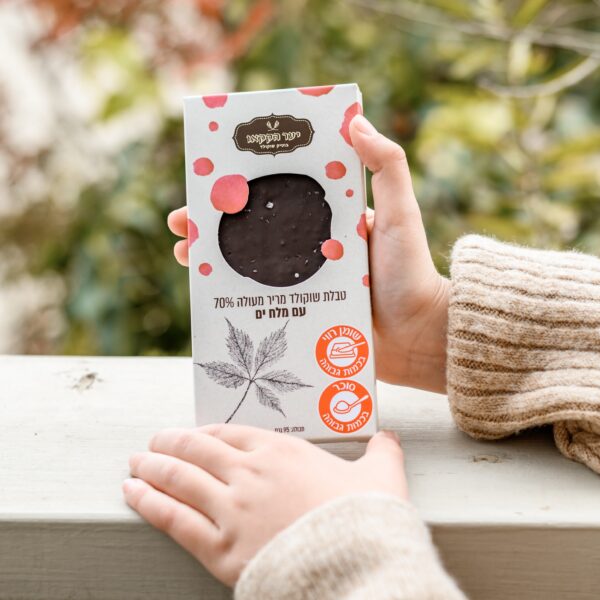 אדם בסוודר בז' מצולע אוחז בקופסת טבלה מריר מלח של שוקולד מריר עם מלח ים וטקסט בעברית, עם אייקונים כתומים, עיצוב עלים ושוקולד לבן. עלווה ירוקה וכתום מטושטשת צובעת את הרקע.