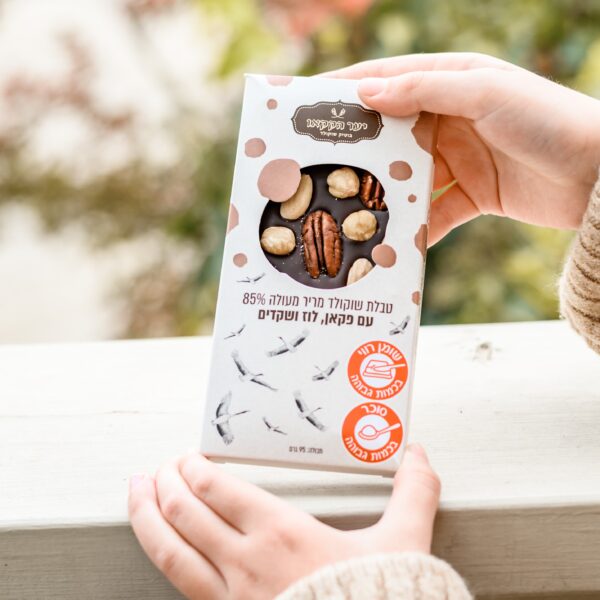 אדם מחזיק טבלה "מריר עם אגוזים" בעטיפה לבנה עם כתמים חומים, כיתוב בעברית ושתי תוויות עגולות כתומות, עומד ליד מעקה בהיר כשברקע ירק מטושטש.