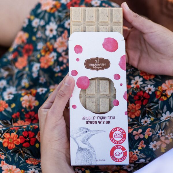 אדם מחזיק חטיף טבלה צאי עם נקודות ורודות, איור של ציפור וטקסט בעברית על הקופסה, כשהוא מונח על ברכיו מעל שמלה עם דוגמת פרחים. האריזה מציעה טעמי צ'אי מסאלה לפינוק ייחודי.