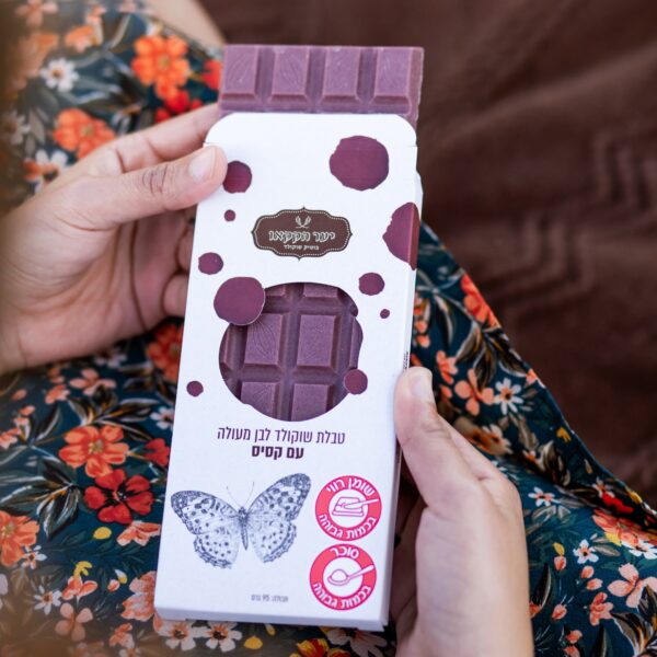 אדם בשמלה פרחונית מחזיק חטיף שוקולד "טבלה קסיס" עם איור פרפר וטקסט בעברית על האריזה. החטיף, כנראה בטעם צ'אי מסאלה, נראה חלקית בתוך הקופסה.