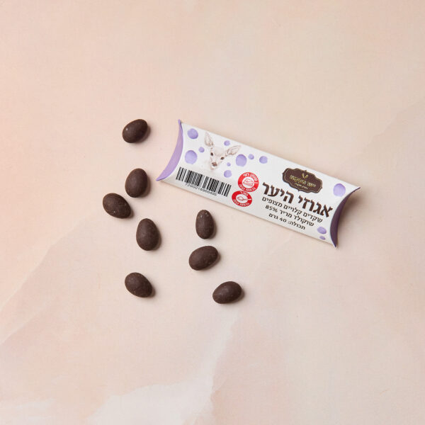 חבילה קטנה עם הכיתוב "IMG_2098" בעברית ומדבקה זהובה מונחת על משטח ורוד בהיר, מוקפת בשמונה שקדים מצופים שוקולד מריר 85% בצורת אליפסה הפזורים סביבה.